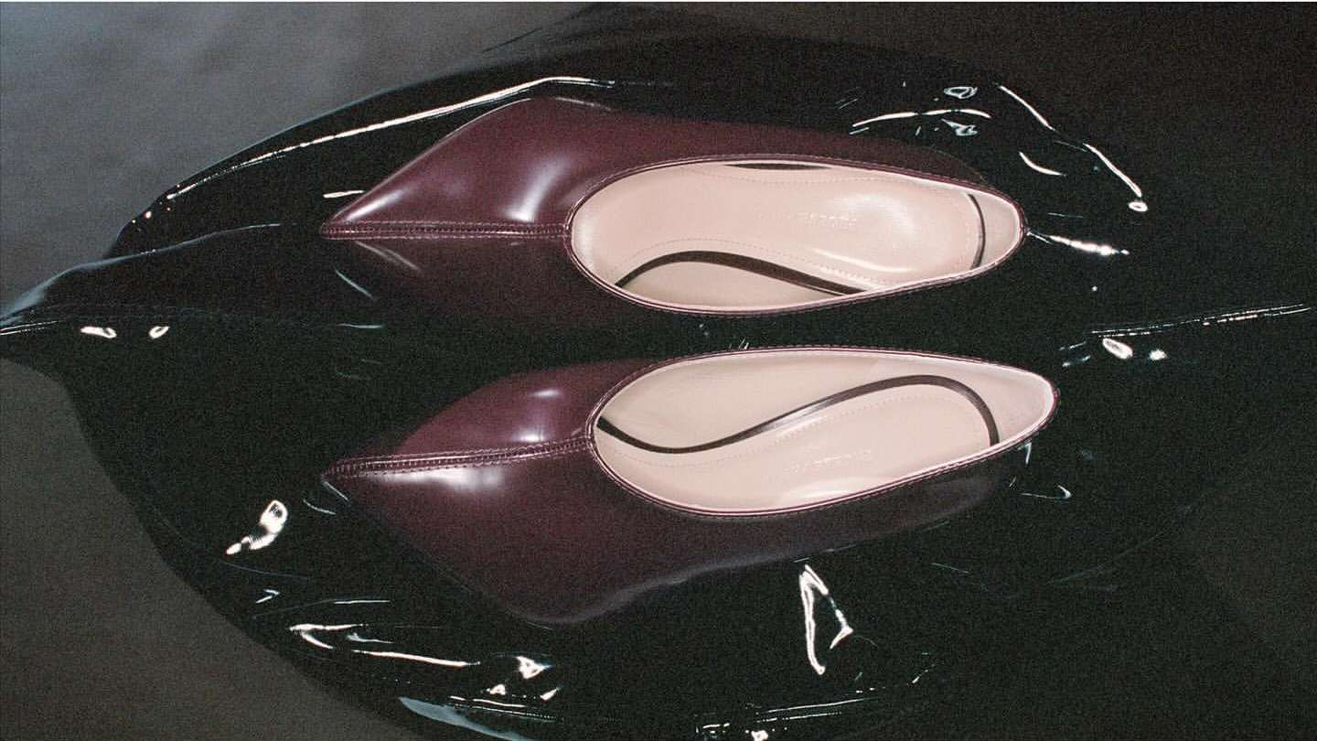 A pair of burgundy leather point-toe flats sit on a glossy black surface in a product shot highlighting the shoe's design and material.