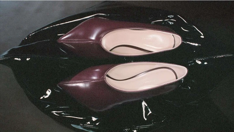 A pair of burgundy leather point-toe flats sit on a glossy black surface in a product shot highlighting the shoe's design and material.
