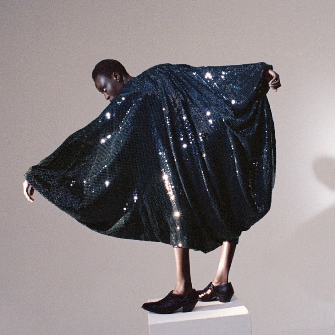 Fashion shot of a Black model in a shimmering dark green dress, posing on a white block against a light gray background.