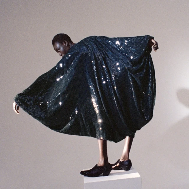 Fashion shot of a Black model in a shimmering dark green dress, posing on a white block against a light gray background.