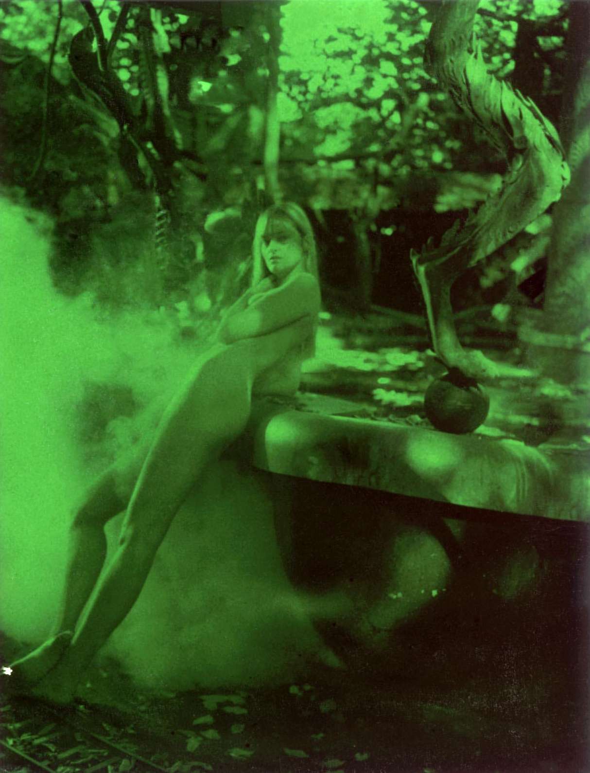Nude woman with long blonde hair rests on a stone structure in a forest setting, highlighted by green light and soft shadows.