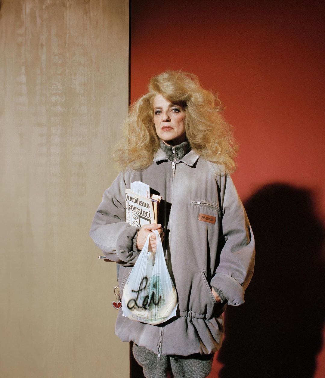 Adult woman with blonde hair and gray tracksuit stands indoors holding newspapers and a shopping bag in a color-blocked setting.