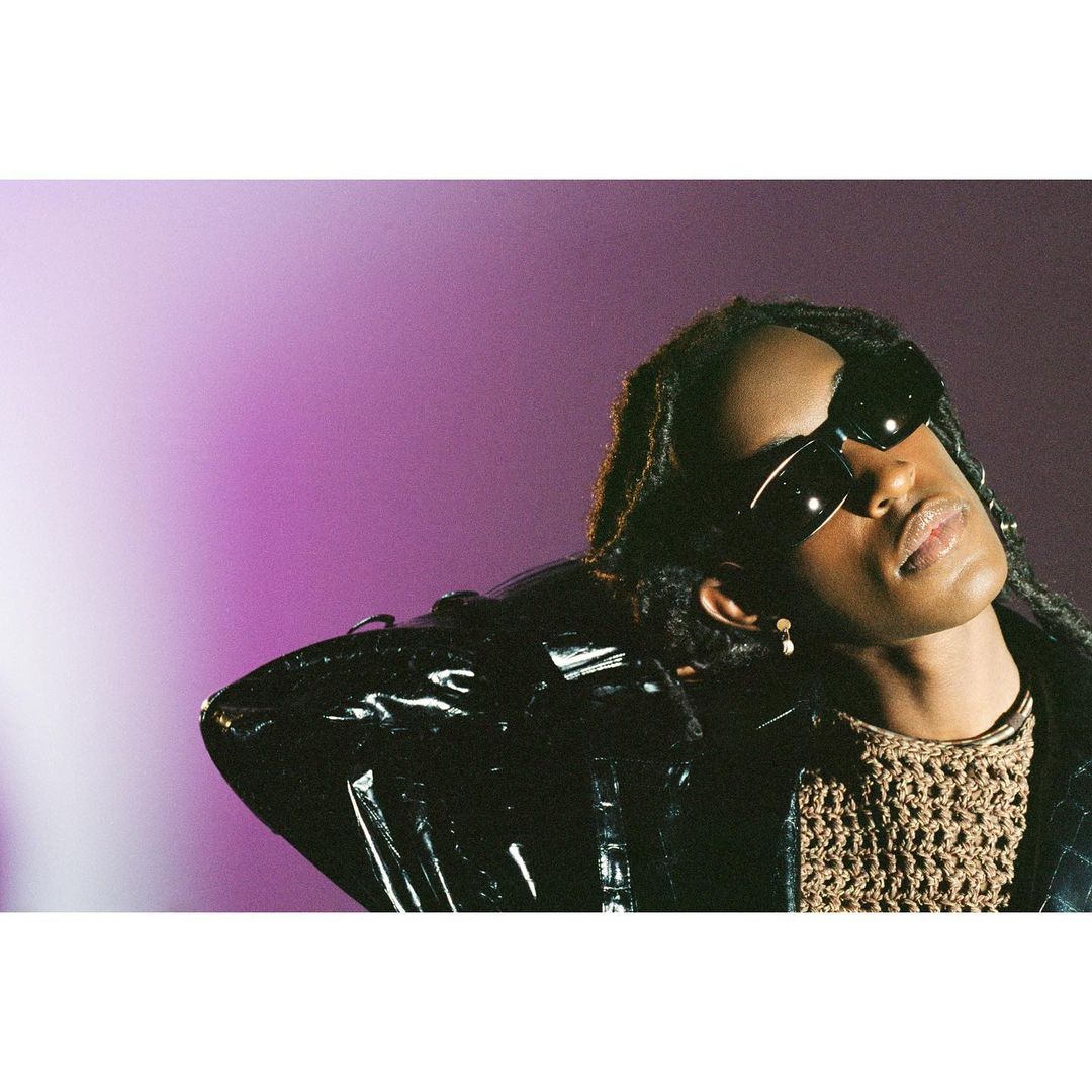 Portrait of a person wearing black sunglasses and a shiny patent jacket against a purple gradient background