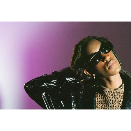 Portrait of a person wearing black sunglasses and a shiny patent jacket against a purple gradient background