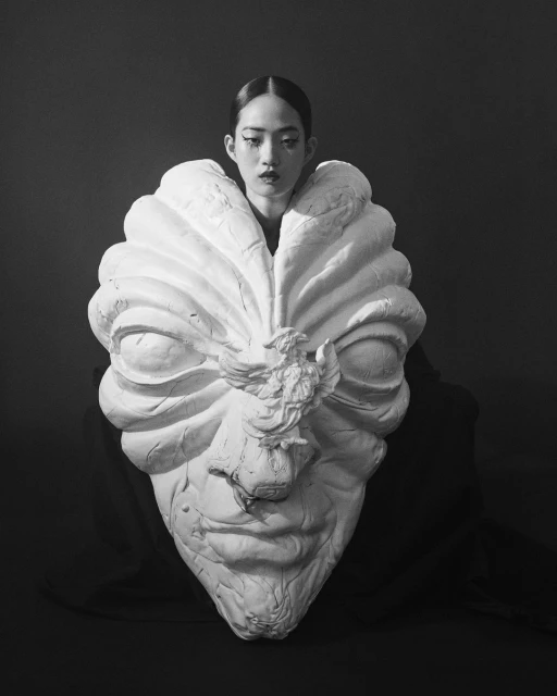 Artistic portrait of a woman with elaborate makeup and an oversized sculpted mask with a regal bird element, black and white tones.