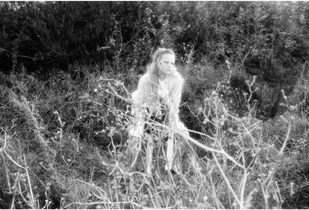 Monochrome portrait of a fair-skinned woman in a light-colored outfit standing amongst tall grass and foliage in an outdoor setting.