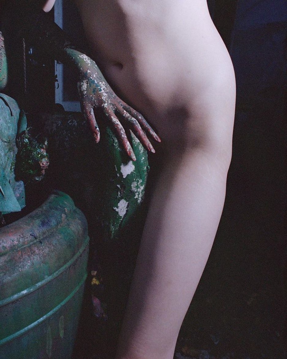 Nude art model posing near a paint-covered statue. The composition features muted color palette, soft lighting, and textured surfaces.