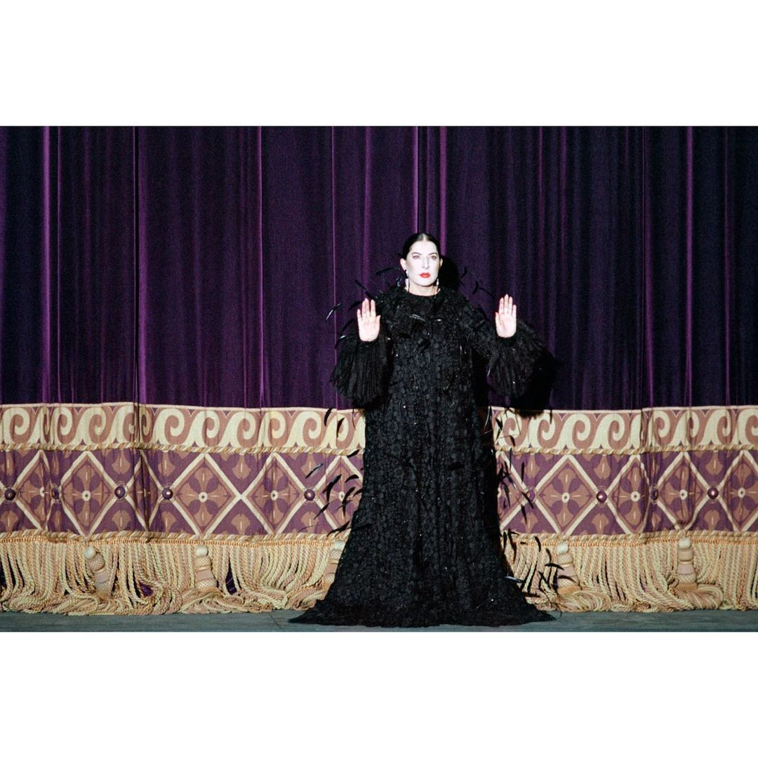 Elegant woman in long black dress with feathers standing on stage in front of purple velvet curtain and ornate gold valance.