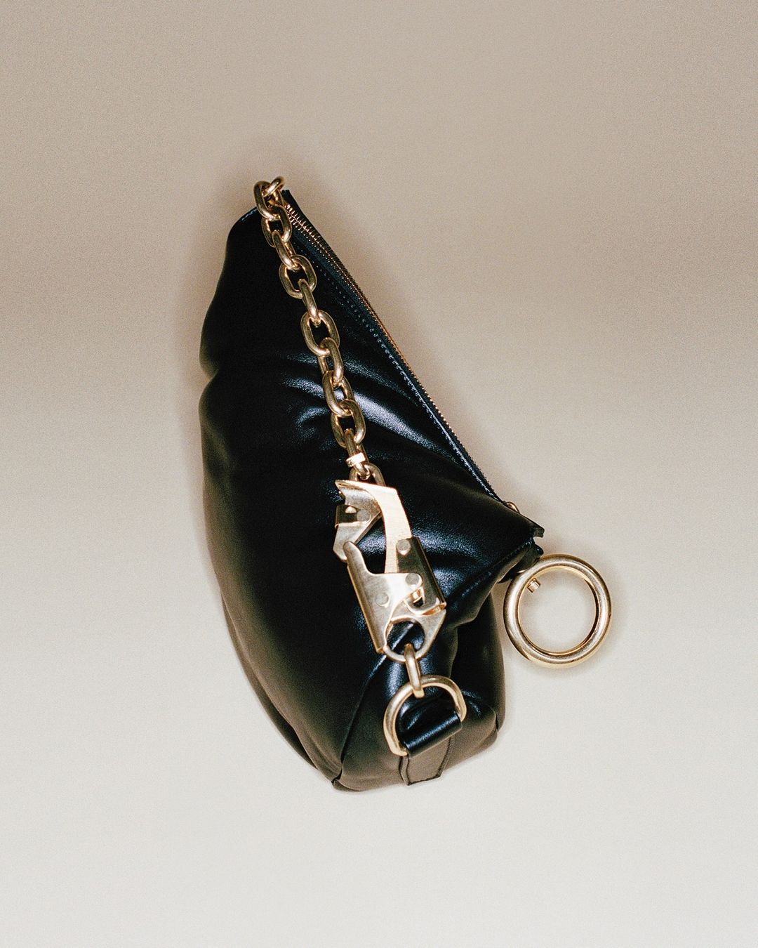 Padded black clutch bag with a gold chain strap, carabiner detail, and gold ring accent, set against a neutral beige background.