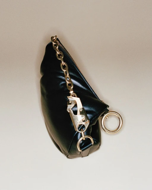 Padded black clutch bag with a gold chain strap, carabiner detail, and gold ring accent, set against a neutral beige background.