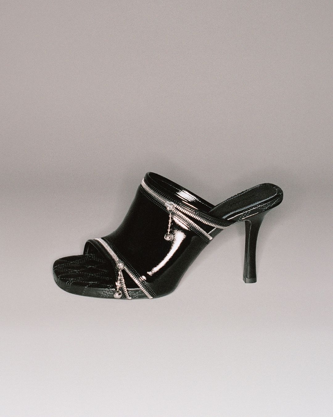 Black patent leather heeled mule with silver zipper detailing on a grey background. The heel is thin and high.  