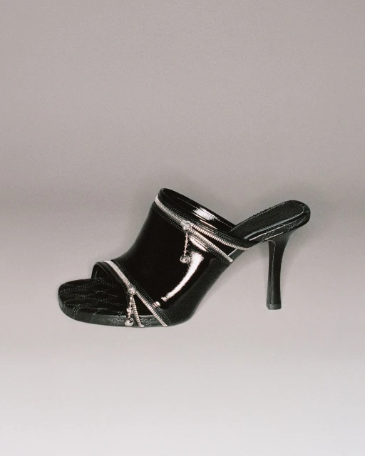 Black patent leather heeled mule with silver zipper detailing on a grey background. The heel is thin and high.  