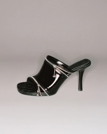 Black patent leather heeled mule with silver zipper detailing on a grey background. The heel is thin and high.  
