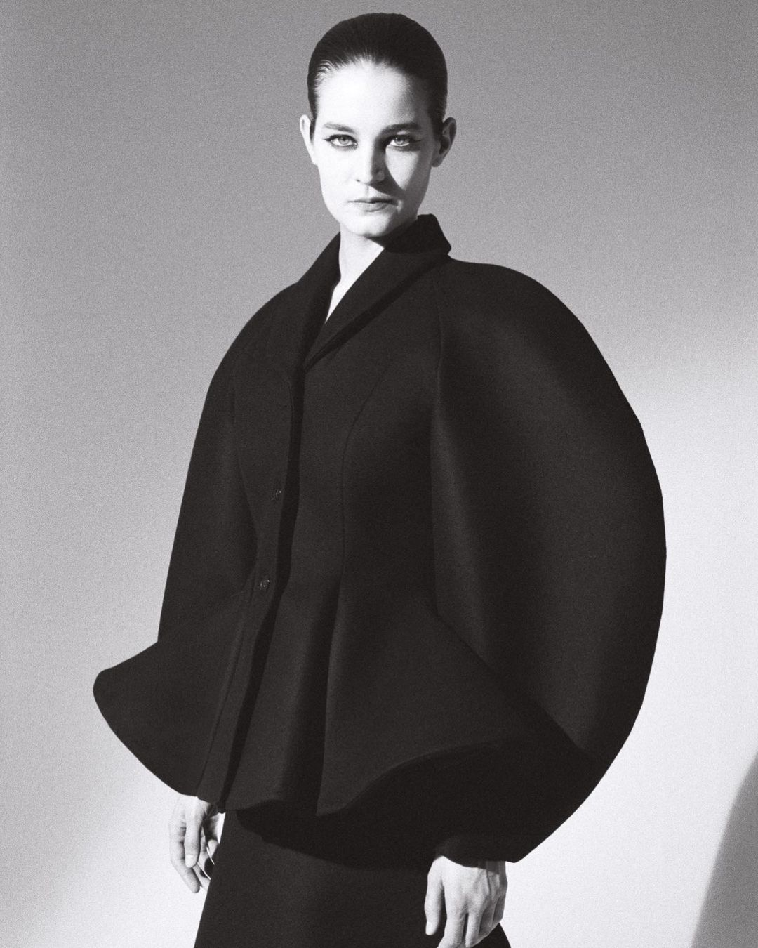 Woman in dramatic black peplum jacket poses against a neutral backdrop in a fashion editorial style studio portrait.
