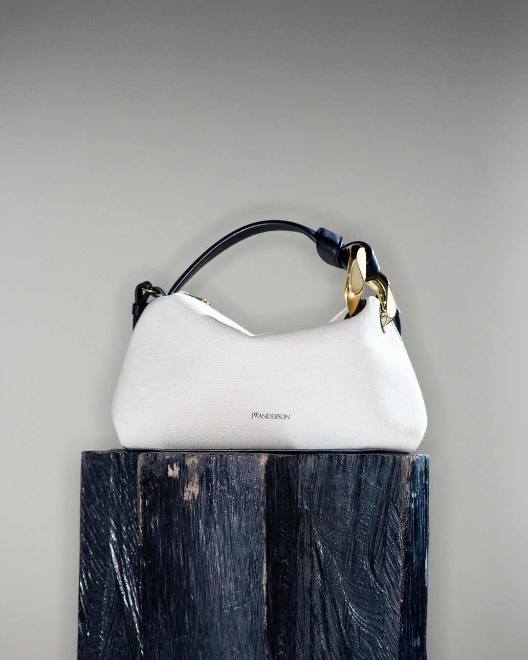 Fashion still life: a luxury white JW Anderson handbag with a black handle and gold hardware on a dark blue wood pedestal.