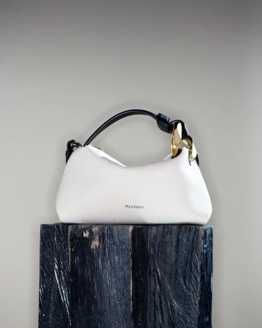 Fashion still life: a luxury white JW Anderson handbag with a black handle and gold hardware on a dark blue wood pedestal.