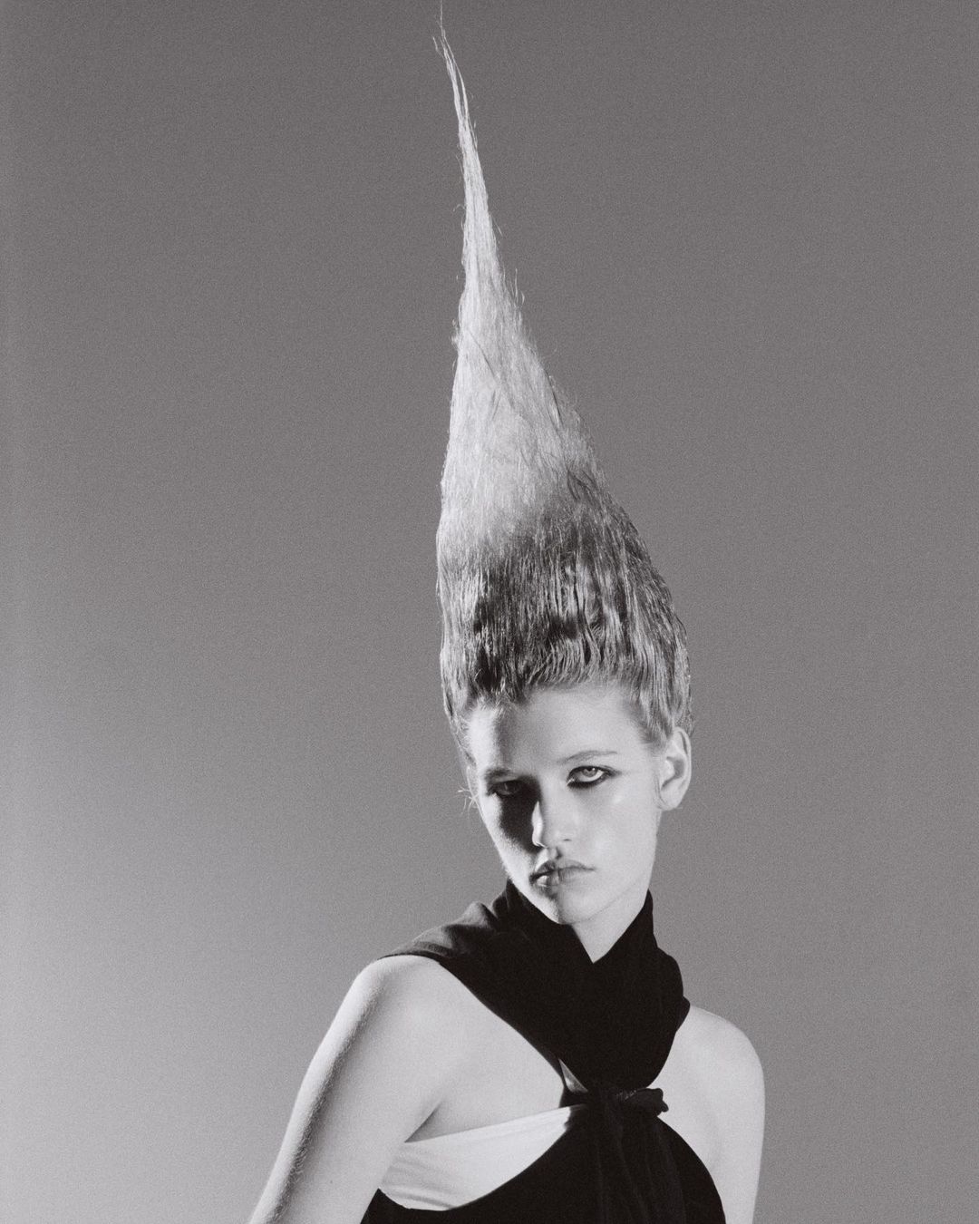 Black and white portrait of a young woman with avant-garde, vertically styled hair and black dress, set against a grey backdrop.