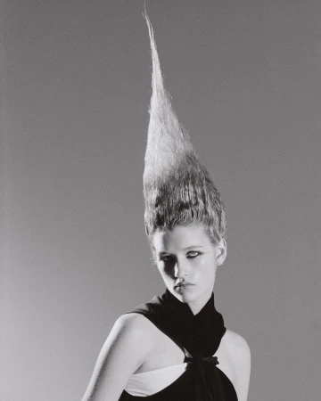 Black and white portrait of a young woman with avant-garde, vertically styled hair and black dress, set against a grey backdrop.