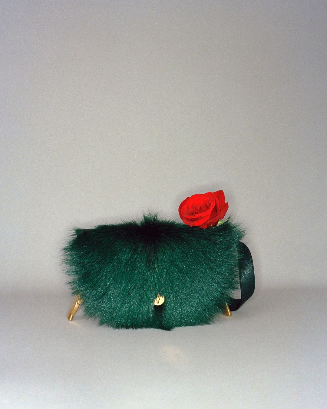 Green faux fur handbag with golden legs and a red rose on a light grey background in a studio setting, product shot.