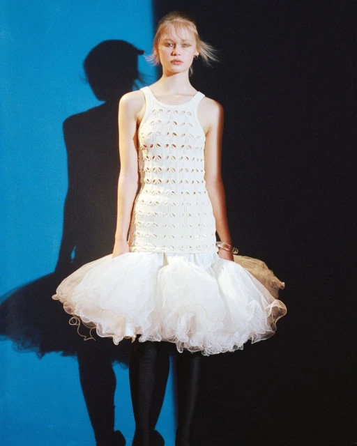 A slender, light-skinned woman models a modern sleeveless top and tutu-style skirt. The image is composed against a split blue and black backdrop.