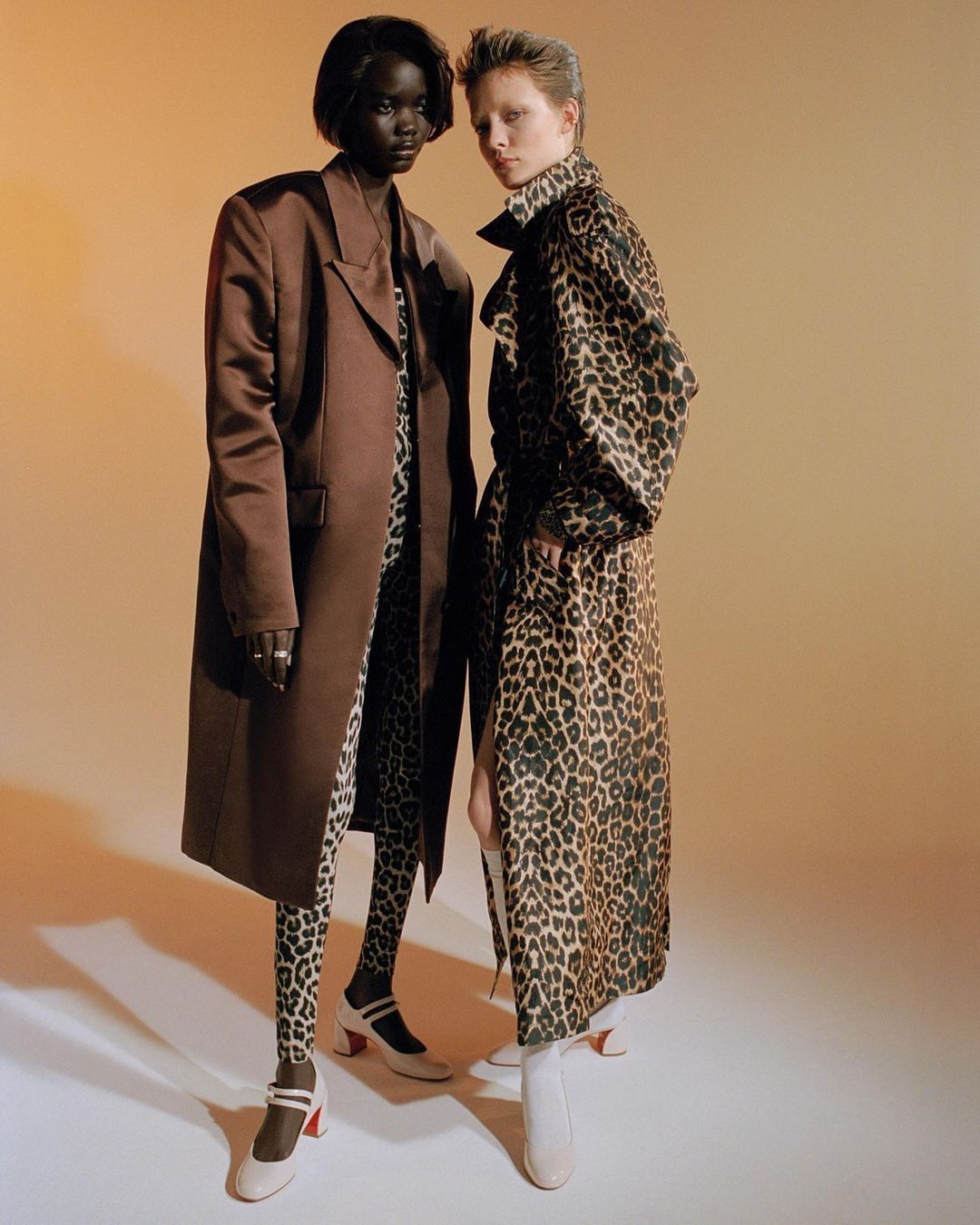 Two women pose in a studio wearing coordinated outfits: one in a brown coat and leopard print, the other in a leopard print coat.
