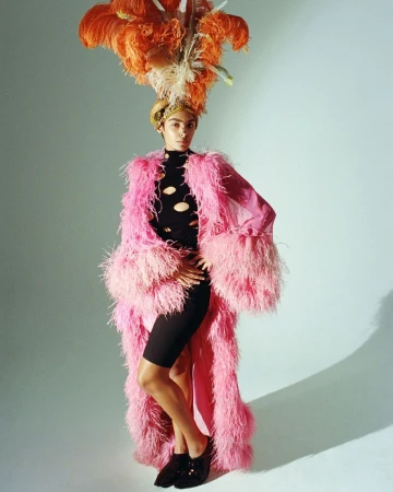 Adult poses in a photo studio wearing a long, pink feather coat, black bike shorts, a black cut-out top, and a large, orange feathered hat.