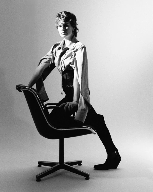 Black and white studio portrait of a model with short hair wearing a blouse and corset, seated on a molded plastic chair.