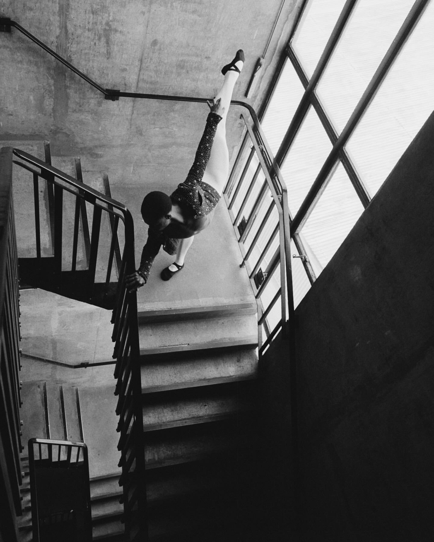 Black and white photo of a woman in a patterned top balancing on a staircase with one leg extended vertically toward a window.