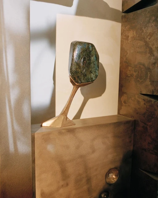 Close-up shot of a blue-green labradorite stone sculpture on a metallic pedestal, set against a textured neutral-toned wall.