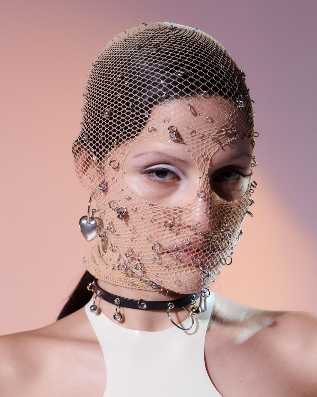 Close-up of an adult woman wearing a net face covering adorned with metallic embellishments, paired with a vinyl choker and heart earring against a gradient background.
