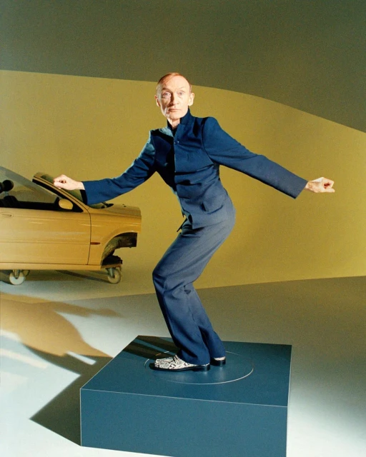 Elderly man in a blue suit strikes a pose on a blue cube in a studio setting, with a yellow deconstructed car in the background.