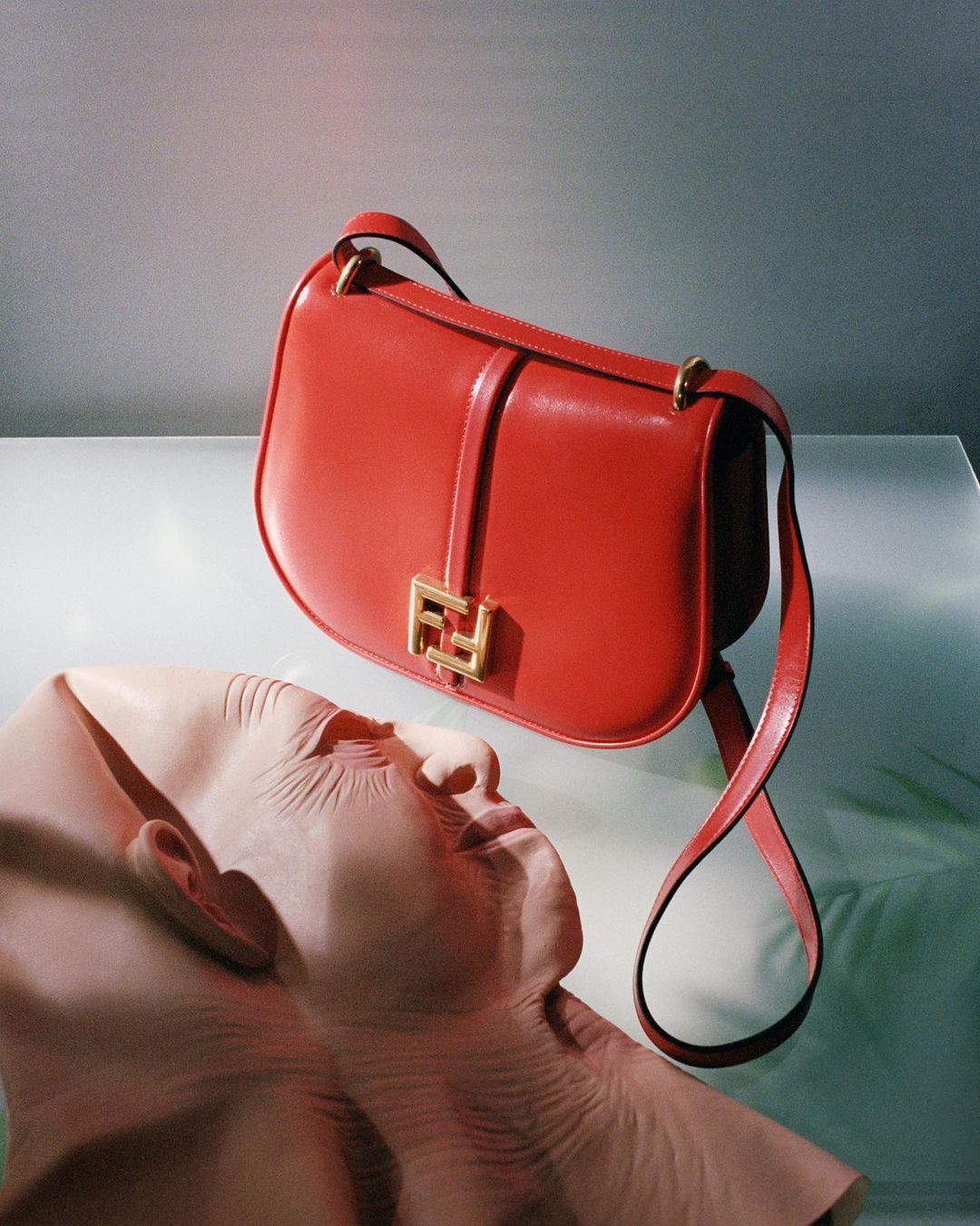 Red Fendi handbag with gold logo hardware on a reflective surface, accompanied by a light pink mannequin head displaying high fashion.