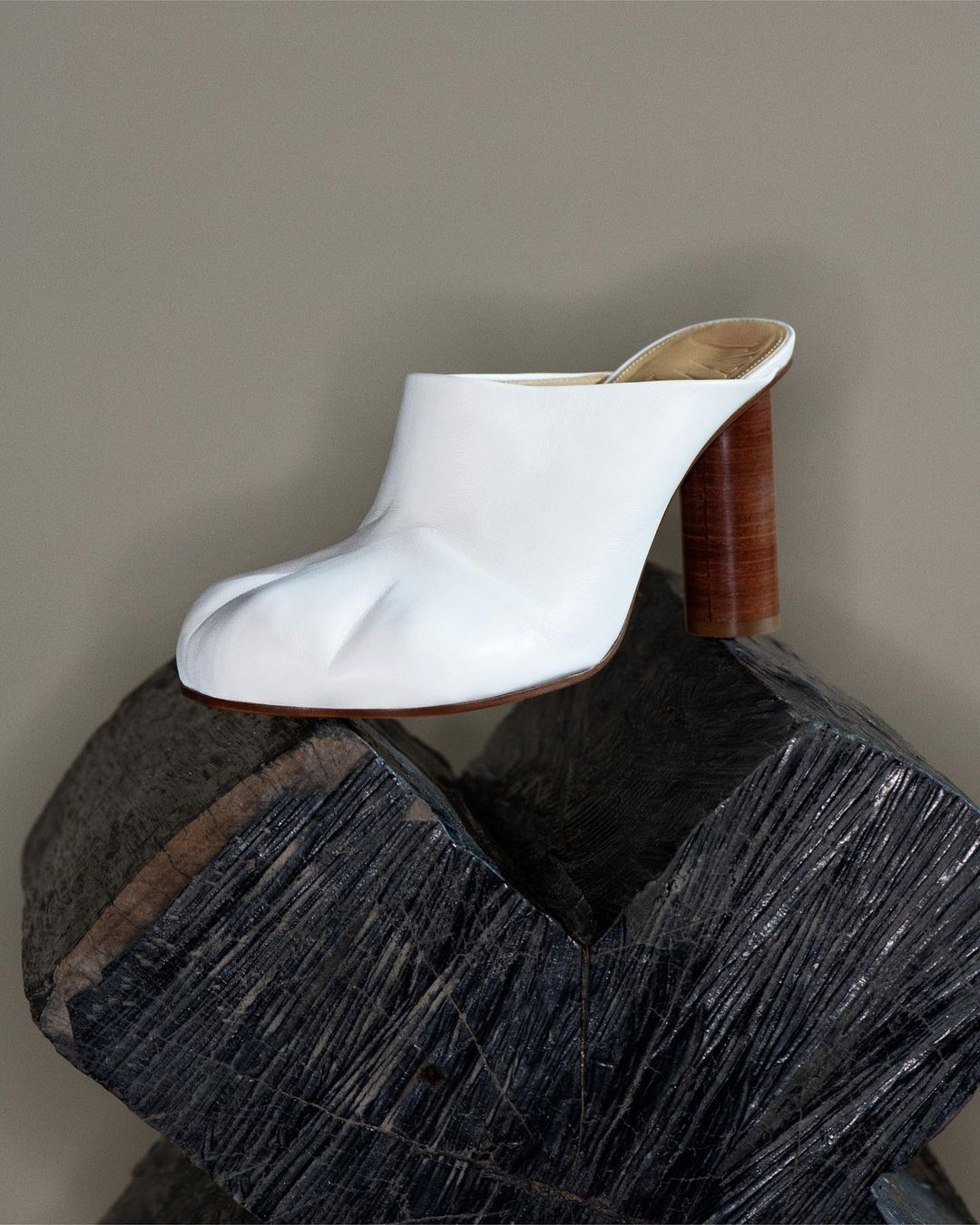 White leather mule shoe with a high wooden heel is placed atop a modern, geometric stone sculpture against a neutral background.