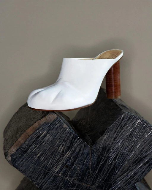 White leather mule shoe with a high wooden heel is placed atop a modern, geometric stone sculpture against a neutral background.