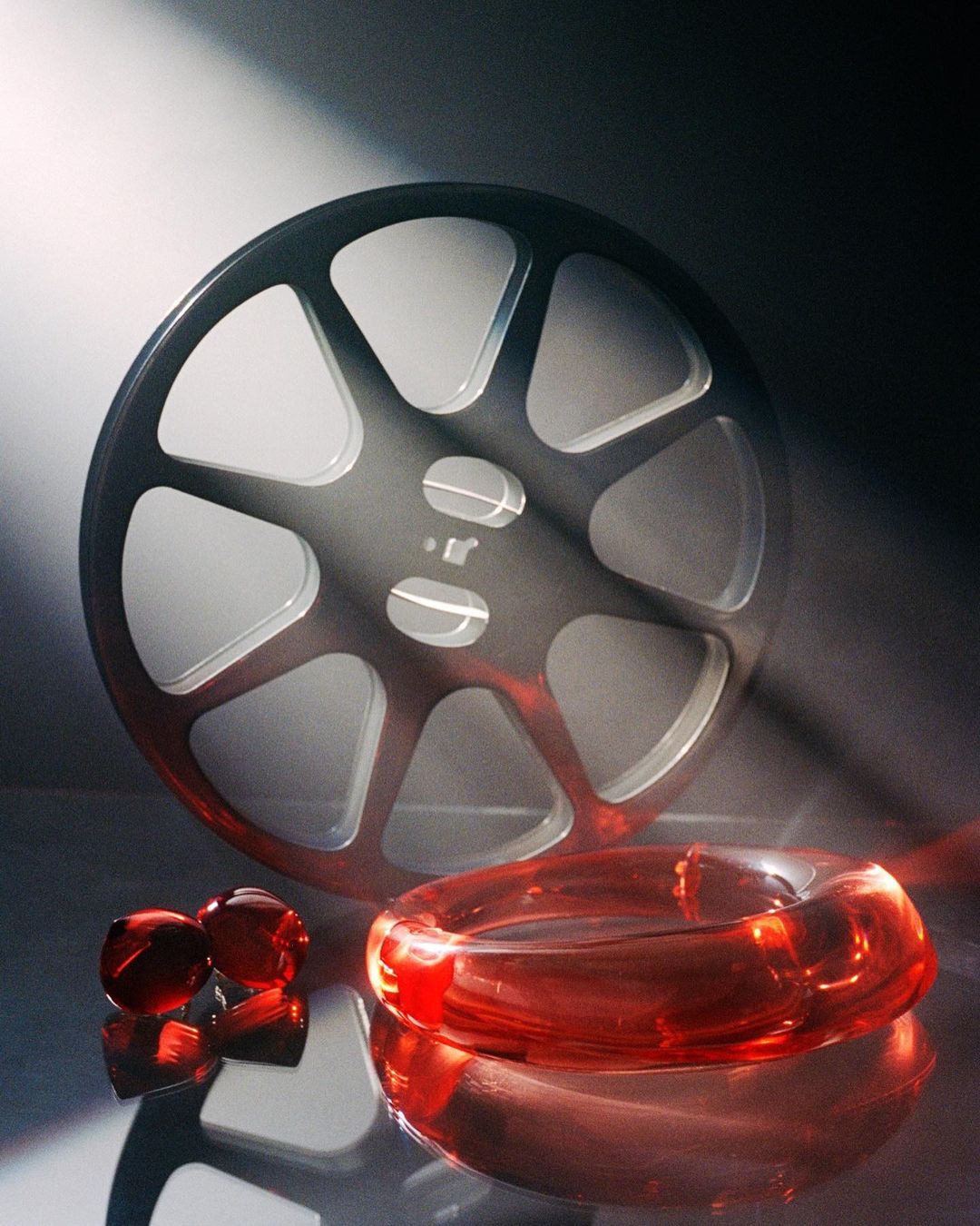 Film reel and red glass objects on a reflective surface are illuminated with a light beam in a studio setting against a dark backdrop.