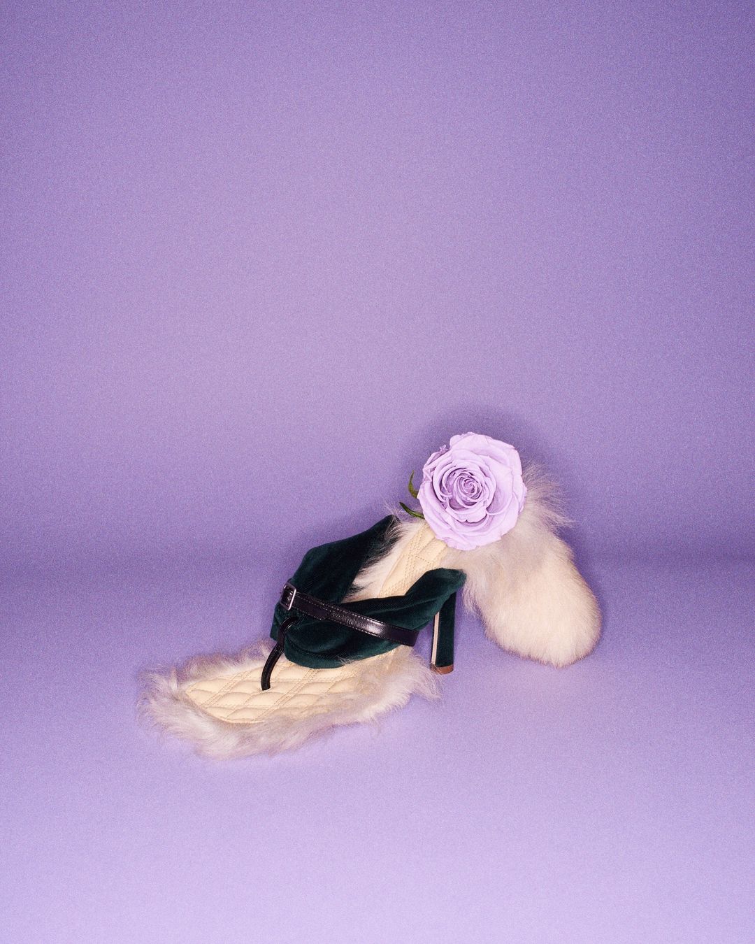 Green high-heeled shoe with a lavender rose and white faux fur accents displayed on a purple backdrop, evoking surreal fashion.