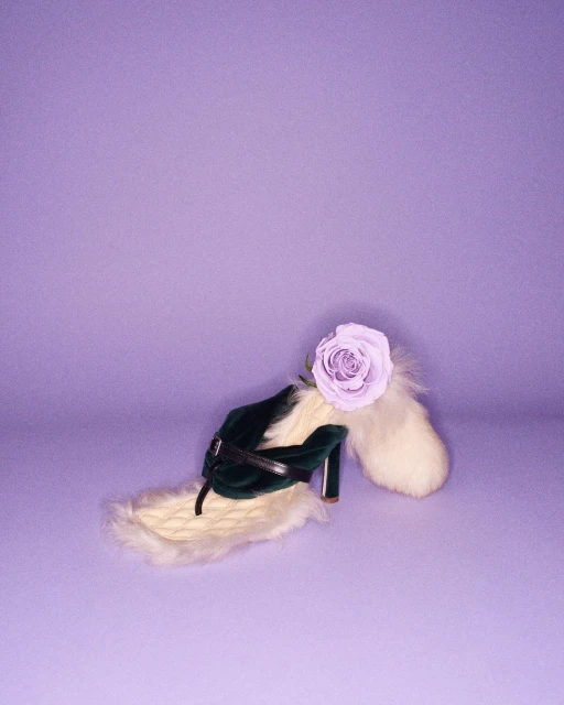 Green high-heeled shoe with a lavender rose and white faux fur accents displayed on a purple backdrop, evoking surreal fashion.