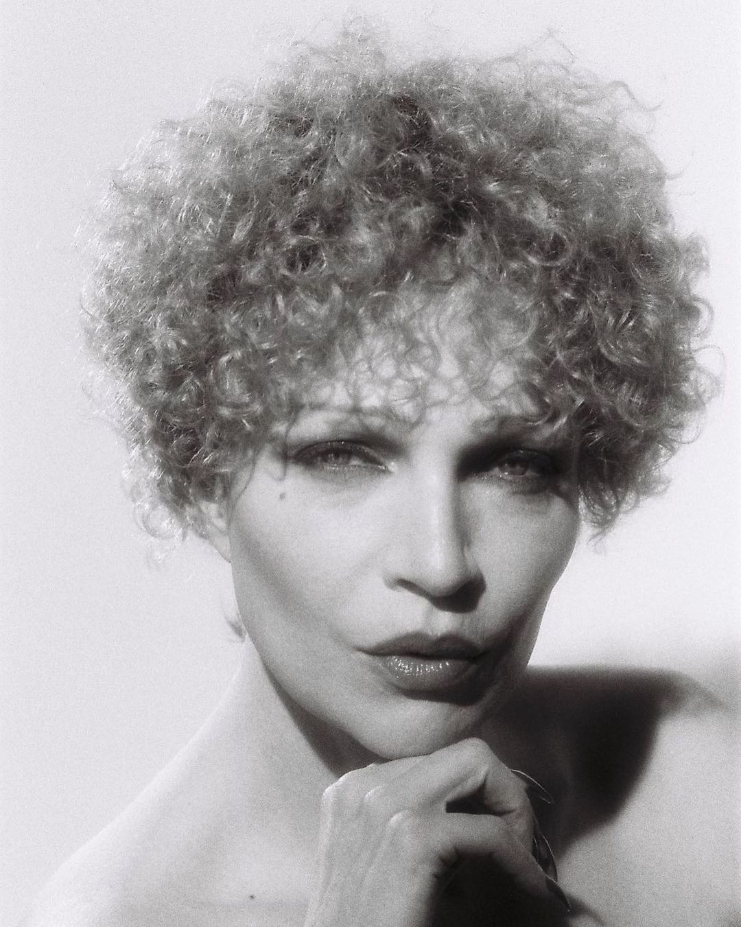 Monochrome portrait of a woman with curly blonde hair, resting her chin on her hand in a studio setting, against a neutral background.