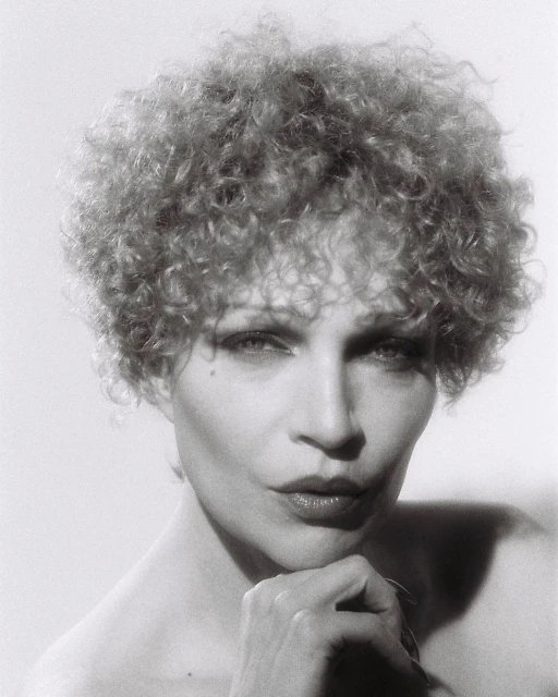 Monochrome portrait of a woman with curly blonde hair, resting her chin on her hand in a studio setting, against a neutral background.