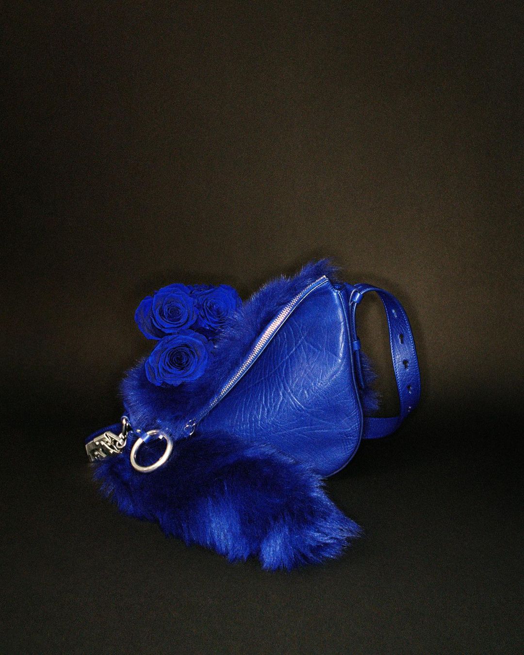 Fashion still life of a blue leather purse trimmed with blue fur and decorated with blue roses against a dark background.