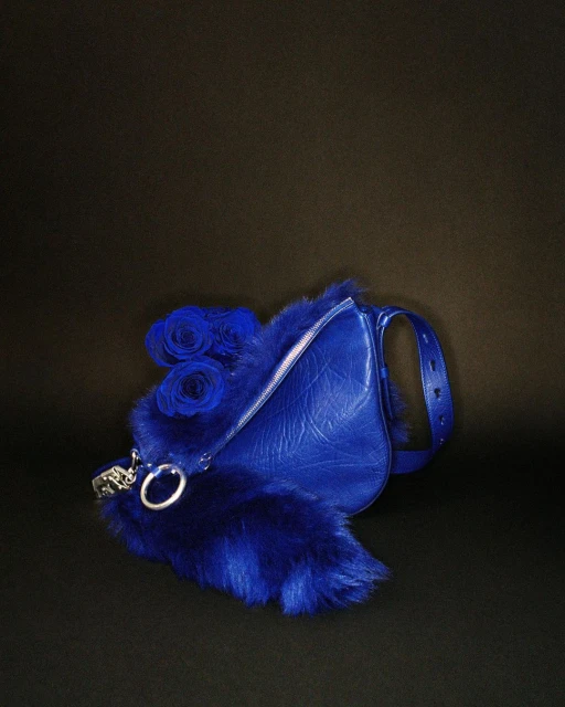 Fashion still life of a blue leather purse trimmed with blue fur and decorated with blue roses against a dark background.