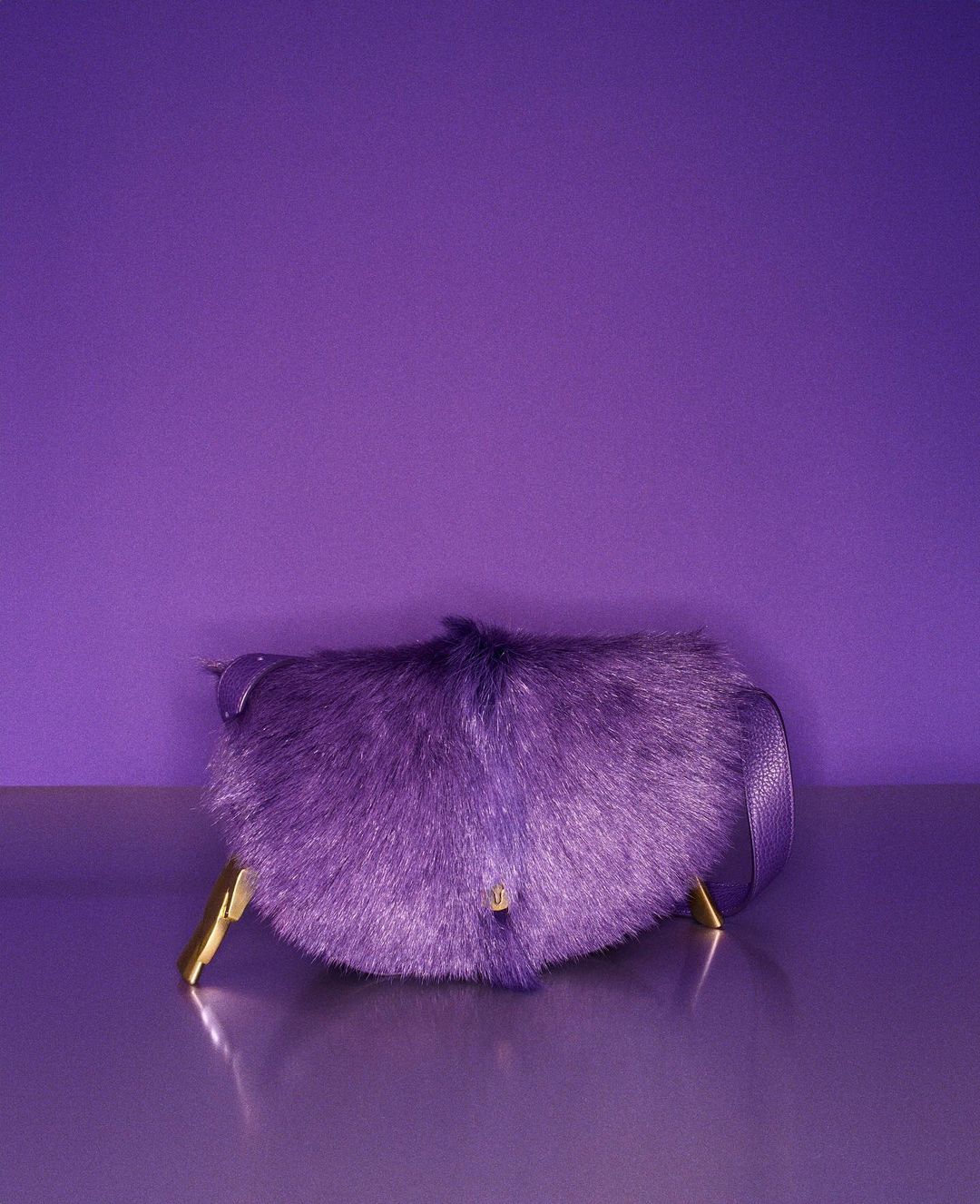 Purple furry handbag with a strap and golden feet in a studio shot against a matching purple background.