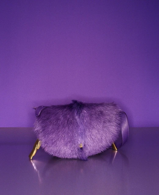Purple furry handbag with a strap and golden feet in a studio shot against a matching purple background.