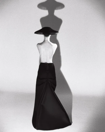 Model from behind wearing a wide-brimmed hat and black skirt against a plain wall with a sharp shadow cast