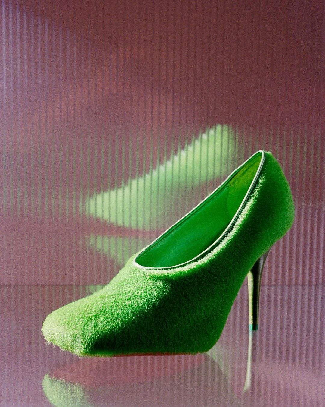 A lime-green, fur-covered, square-toed pump with stiletto heel stands against a glossy pink surface with a vertically ribbed backdrop.