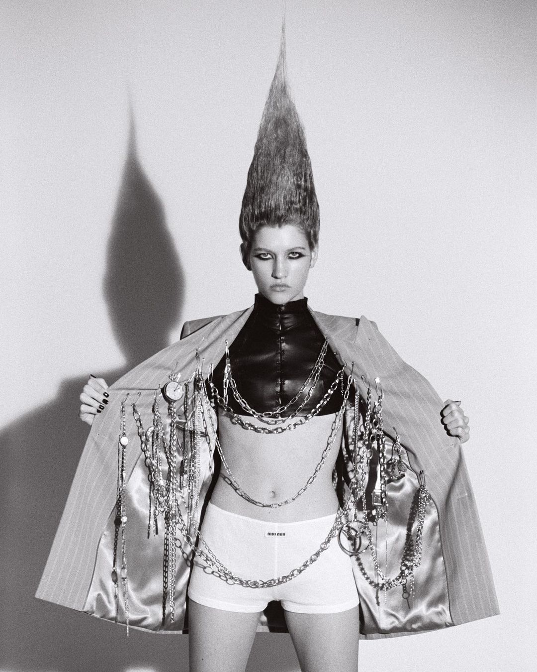 Woman with striking hairstyle wearing pinstripe jacket and chains in a studio. Fashion-forward and bold visual statement, black and white photograph.