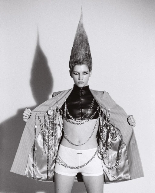Woman with striking hairstyle wearing pinstripe jacket and chains in a studio. Fashion-forward and bold visual statement, black and white photograph.