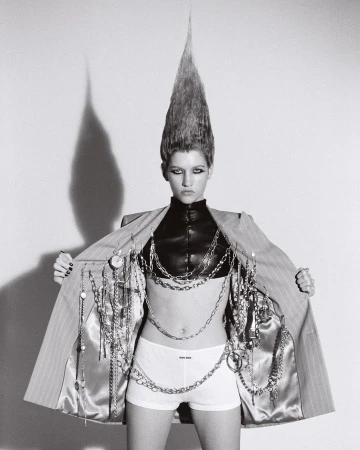 Woman with striking hairstyle wearing pinstripe jacket and chains in a studio. Fashion-forward and bold visual statement, black and white photograph.