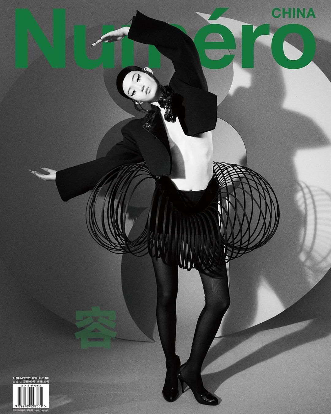 Fashion editorial image features a slender adult woman wearing a black jacket, skirt, and heels on a grey patterned backdrop.