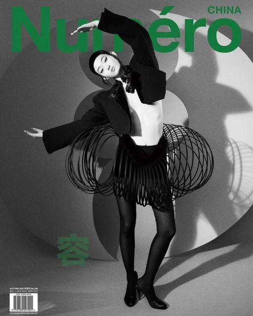 Fashion editorial image features a slender adult woman wearing a black jacket, skirt, and heels on a grey patterned backdrop.